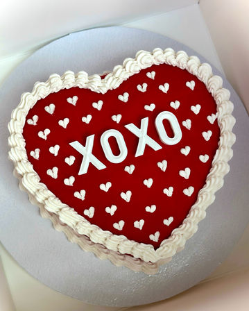Heart-shaped red Valentine’s cake with ruffled white icing border, small piped white hearts and bold 'XOXO' letters on a silver cake board — festive romantic dessert.