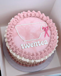 Pink ruffle-frosted layered celebration cake in a takeaway box with pearl-piped teacup outline, white lettered message on top and a small pink bow decoration.