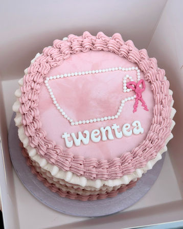 Pink ruffle-frosted layered celebration cake in a takeaway box with pearl-piped teacup outline, white lettered message on top and a small pink bow decoration.