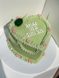 Two-tier heart-shaped pistachio-green cake with ruffled piped icing, pink buttercream roses and pearl accents, topped with a skewered green olive and white lettering reading “bride in a tinibit.”