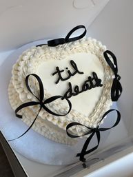 Heart-shaped white buttercream cake with black cursive 'til death', black ribbon bows and pearl accents in a cake box.