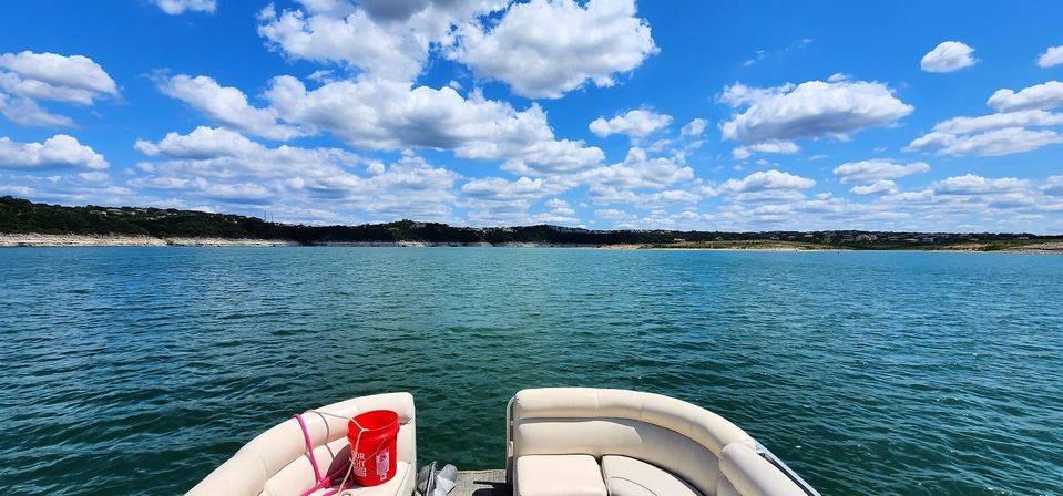 View from a pontoon boat bow with cream seats and a red bucket, looking out over a turquoise lake to a tree-lined shoreline under a bright blue sky with fluffy white clouds
