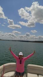 Person in a bright pink shirt and white cap raising both arms aboard a pontoon boat on a sunny lake with sparkling water and fluffy clouds under a clear blue sky.