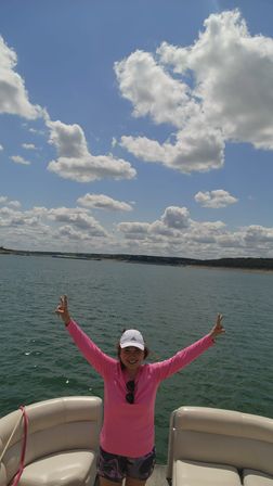 Person in a bright pink shirt and white cap raising both arms aboard a pontoon boat on a sunny lake with sparkling water and fluffy clouds under a clear blue sky.