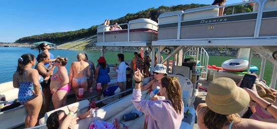 Group of people enjoying a sunny summer pontoon boat party on a green lake, wearing swimsuits, holding drinks with inflatable tubes and a tree-covered shoreline in the background.