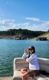 Person relaxing on a pontoon boat on a sunny lake, flashing a peace sign while wearing a cap, long-sleeve shirt and leopard-print shorts with green hills and blue sky in the background