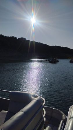 Glittering lake scene with bright sun and rainbow lens flare, sparkling water reflection, pontoon boat seating in the foreground and silhouetted hillside with distant boats