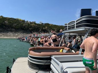 Sun-soaked lake party with dozens of people in swimsuits crowded on linked pontoon boats and a dock near a rocky shoreline and tree‑covered hillside under a clear blue sky.