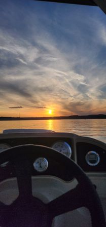Sunset over a calm lake seen from a boat cockpit — steering wheel and dashboard gauges in foreground, golden sun reflecting on rippled water beneath wispy clouds.