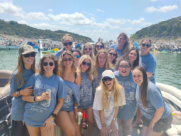 Smiling group of women in matching 'Austin, TX' shirts and sunglasses posing on a crowded summer lake boat with other boats and rocky shoreline under a sunny blue sky.