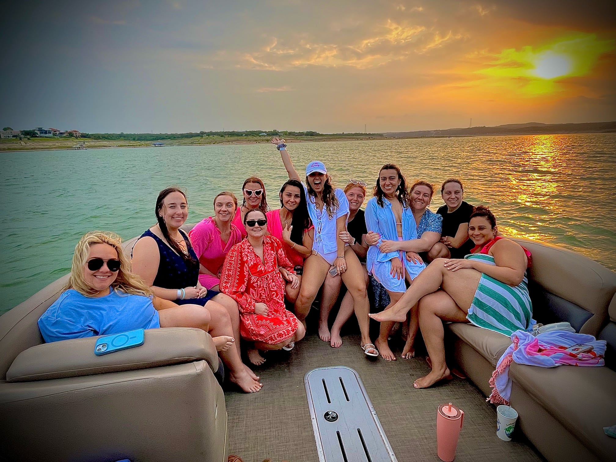 Group of friends on a pontoon boat at sunset on a lake, smiling women in colorful swimwear and cover-ups with the golden sun reflecting on the water