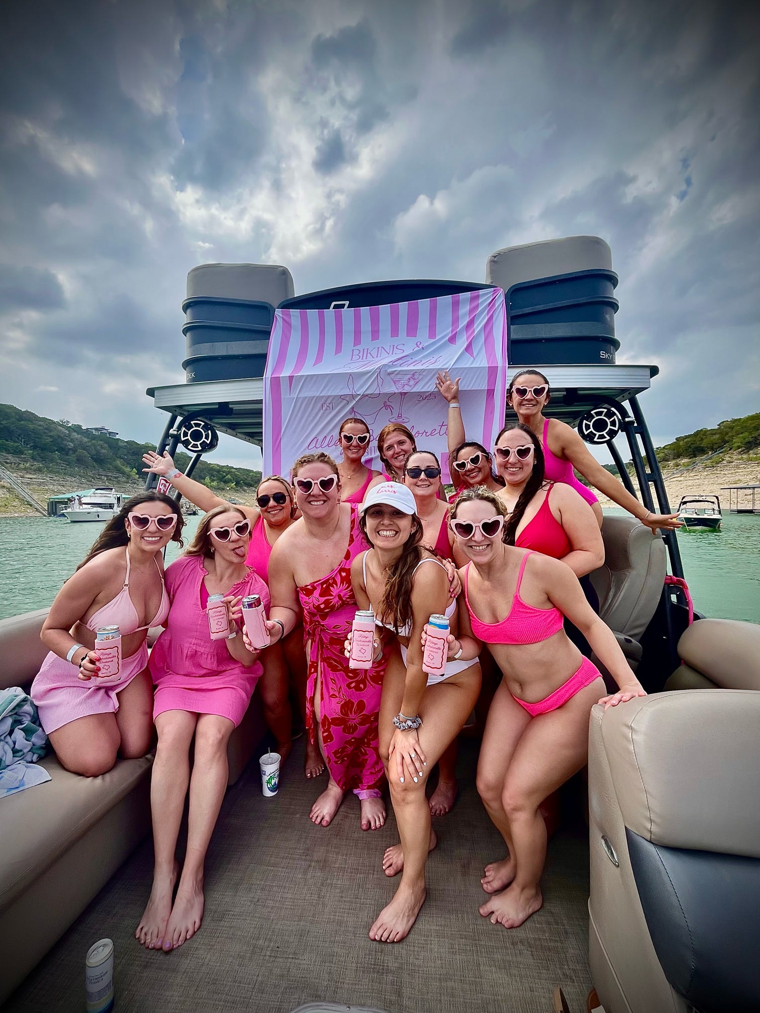 Group of women in pink swimsuits and heart-shaped sunglasses posing and holding drinks on a pontoon boat at a lake in front of a pink-striped party banner under a cloudy sky