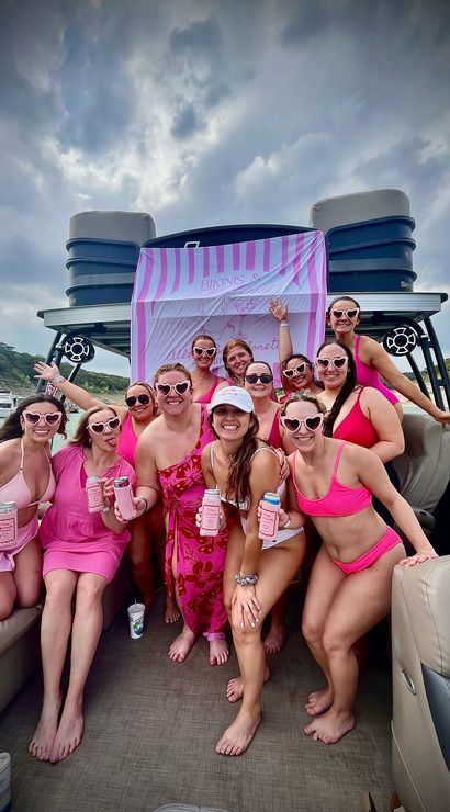 Group of women in pink swimsuits and heart-shaped sunglasses posing and holding drinks on a pontoon boat at a lake in front of a pink-striped party banner under a cloudy sky
