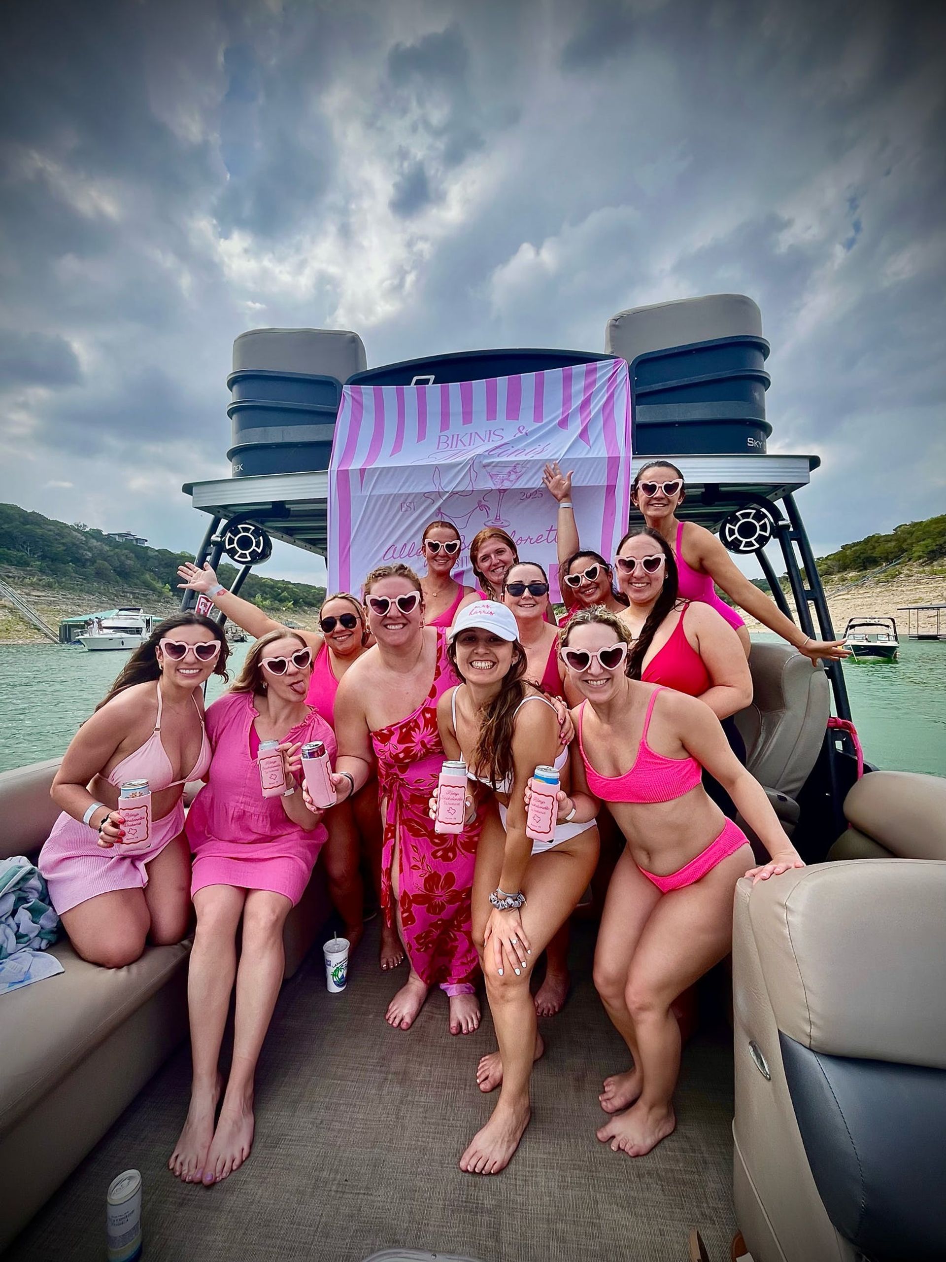 Group of women in pink swimsuits and heart-shaped sunglasses posing and holding drinks on a pontoon boat at a lake in front of a pink-striped party banner under a cloudy sky