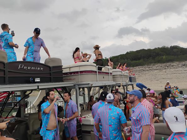 Pontoon boat lake party with people in colorful matching tropical shirts and swimwear socializing and drinking on upper and lower decks near a rocky shoreline and tree-covered hills under an overcast sky.