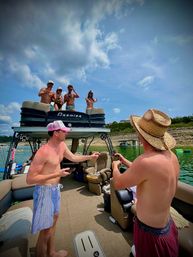 Friends partying on a two-level pontoon boat on a sunny lake in swimwear — straw hat, drinks, rocky shoreline and blue sky