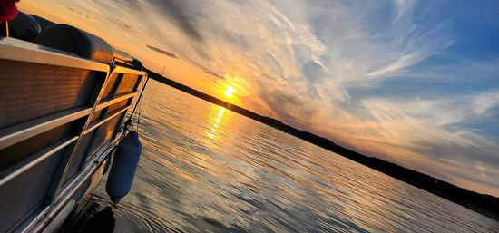 Tilted view from a pontoon boat at sunset on a calm lake, golden sun reflecting on rippling water beneath a colorful, cloud-streaked sky.