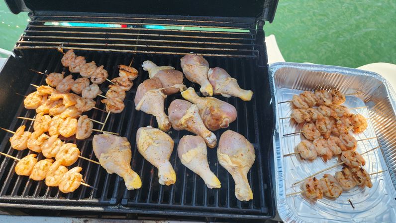 Grill loaded with seasoned shrimp skewers and chicken drumsticks, extra shrimp in a foil tray beside the grill at a sunny waterfront barbecue