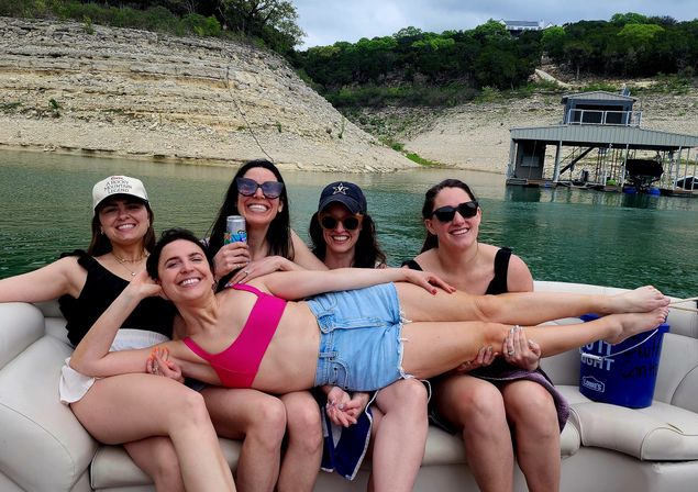 Five friends relaxing on a pontoon boat in a scenic lake cove with limestone cliffs; one woman in a pink bikini top and denim shorts is lying across the group while others laugh, wearing sunglasses and hats and holding a canned drink.