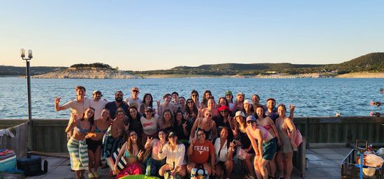 Smiling group of young adults posing on a wooden dock by a lake at golden hour, wearing swimsuits and summer gear with rolling hills in the background