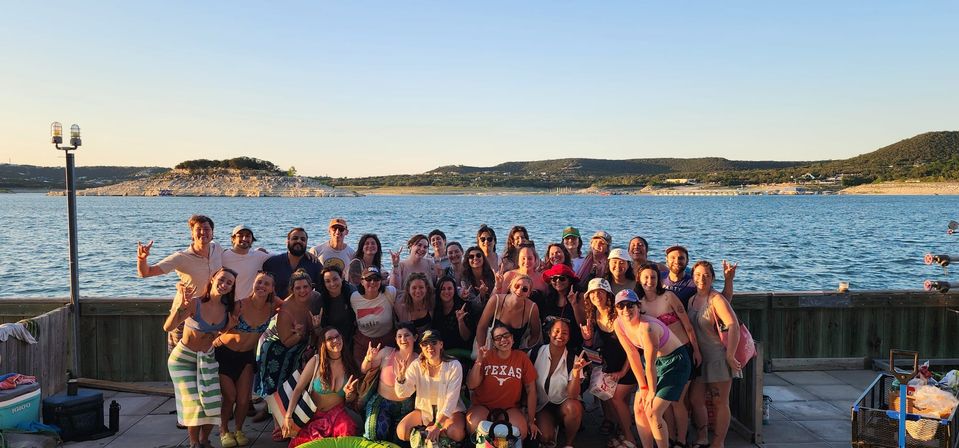 Smiling group of young adults posing on a wooden dock by a lake at golden hour, wearing swimsuits and summer gear with rolling hills in the background