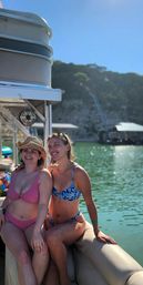 Two friends smiling on a pontoon boat at a sunny lake — one in a pink bikini and straw hat, the other in a blue floral bikini with tree-covered cliffs and docks in the background.