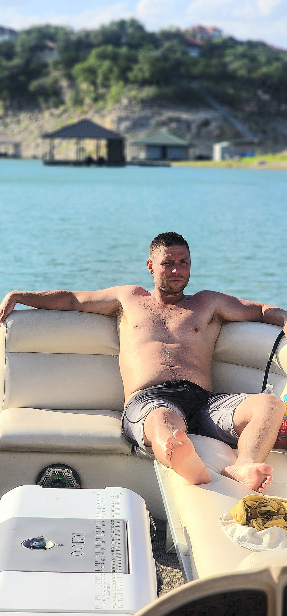 Shirtless man in swim trunks lounging on a pontoon boat with a cooler, lake water and a boathouse-lined shoreline in the background.