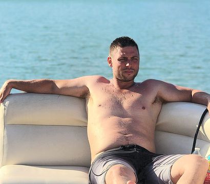 Shirtless man in swim trunks lounging on a pontoon boat with a cooler, lake water and a boathouse-lined shoreline in the background.
