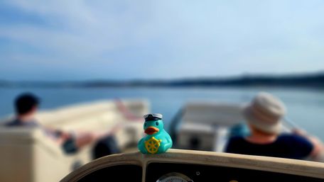 Cheerful turquoise rubber duck wearing a captain’s hat and holding a yellow ship wheel perched on a boat railing, with a calm lake and two blurred passengers in the background