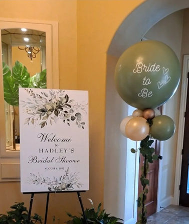 Indoor foyer bridal shower display with a floral welcome sign on an easel, potted greenery, and a tall green-and-gold "Bride to Be" balloon column — elegant neutral decor for a bridal shower entryway.