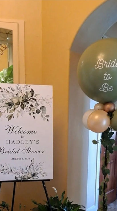 Indoor foyer bridal shower display with a floral welcome sign on an easel, potted greenery, and a tall green-and-gold "Bride to Be" balloon column — elegant neutral decor for a bridal shower entryway.