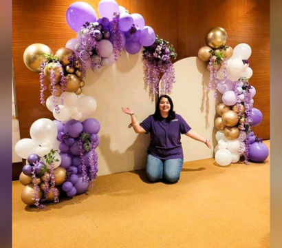 Purple and gold balloon arch with cascading lavender florals framing a white backdrop, person kneeling and presenting the garland in a carpeted indoor event space — stylish balloon photo backdrop for parties and weddings.