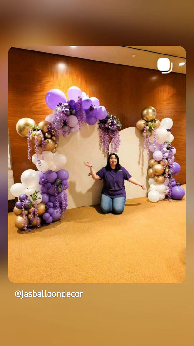 Purple and gold balloon arch with cascading lavender florals framing a white backdrop, person kneeling and presenting the garland in a carpeted indoor event space — stylish balloon photo backdrop for parties and weddings.
