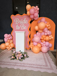 Pink and orange bridal shower backdrop with "Bride to Be" sign, clustered peach and pink balloon garlands, white podium and floral arrangement on a blush patterned rug