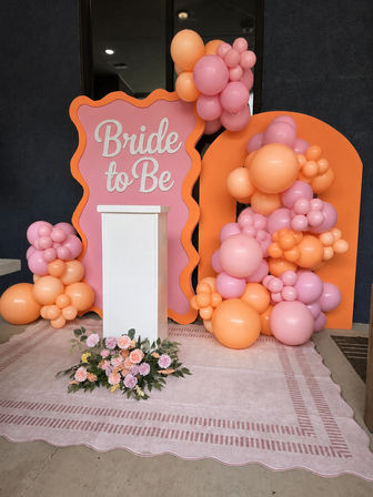 Pink and orange bridal shower backdrop with "Bride to Be" sign, clustered peach and pink balloon garlands, white podium and floral arrangement on a blush patterned rug