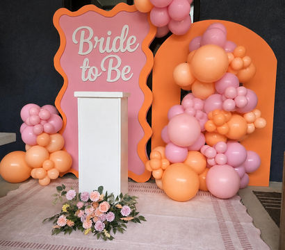 Pink and orange bridal shower backdrop with "Bride to Be" sign, clustered peach and pink balloon garlands, white podium and floral arrangement on a blush patterned rug