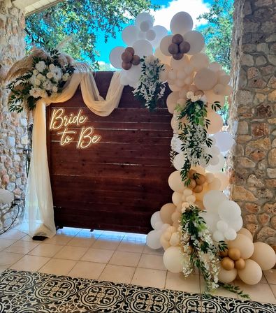 Boho neutral bridal shower backdrop on a stone patio: dark wood panel with neon "Bride to Be" sign, beige and white balloon garland with gold accents, cascading greenery, white florals and sheer drape over patterned tile flooring.