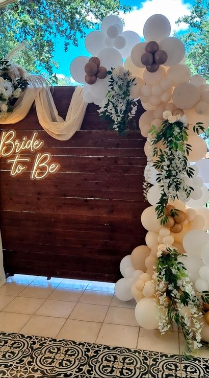 Boho neutral bridal shower backdrop on a stone patio: dark wood panel with neon "Bride to Be" sign, beige and white balloon garland with gold accents, cascading greenery, white florals and sheer drape over patterned tile flooring.