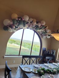 Pastel balloon garland draped over a large arched window overlooking green hills, above a dining table set with floral china, glassware and eucalyptus centerpiece.