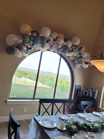 Pastel balloon garland draped over a large arched window overlooking green hills, above a dining table set with floral china, glassware and eucalyptus centerpiece.