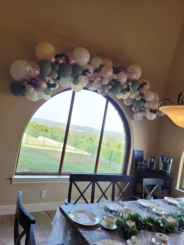 Pastel balloon garland draped over a large arched window overlooking green hills, above a dining table set with floral china, glassware and eucalyptus centerpiece.