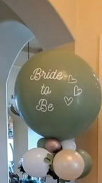 Sage-green “Bride to Be” jumbo balloon column with cream, gold and green clusters, eucalyptus garland and potted plants in an indoor entryway bridal shower display