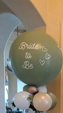 Sage-green “Bride to Be” jumbo balloon column with cream, gold and green clusters, eucalyptus garland and potted plants in an indoor entryway bridal shower display