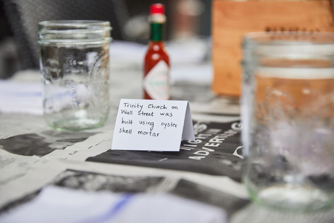 Folded handwritten card on a newspaper-covered table noting a historic church was built with oyster shell mortar, flanked by two glass mason jars and a small hot sauce bottle in the background.