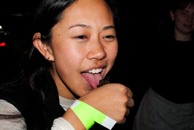 Person at a nighttime food-tasting event licking a spoonful of dark caviar, wearing a neon green wristband