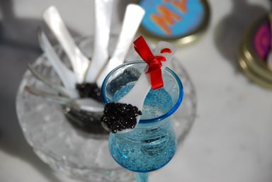Mother-of-pearl tasting spoon topped with black caviar and tied with a red ribbon, perched on textured blue glassware at a gourmet caviar tasting with extra white spoons on ice in the background.