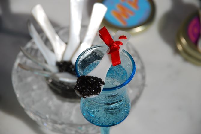 Mother-of-pearl tasting spoon topped with black caviar and tied with a red ribbon, perched on textured blue glassware at a gourmet caviar tasting with extra white spoons on ice in the background.