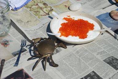 Bright orange salmon roe piled on creamy white sauce in a bowl set on newspaper at an outdoor seafood picnic, with a mason jar, small blue fork and decorative metal crab nearby.