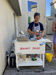 Person in apron behind a white "Market Price" seafood cart on a wooden deck preparing oysters on ice with lemon wedges, hot sauce bottles and a yellow bucket nearby.