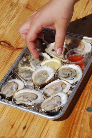 Hand reaching for fresh shucked oysters on the half shell in a metal tray with a lemon wedge and dipping sauce on a sunny wooden table